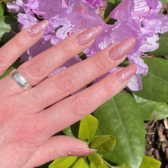 Glänzende rosa Nagelfolien im Sonnenlicht, mit zarten lila Blumen im Hintergrund.