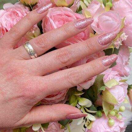 Detailaufnahme von LIGHT PINK glitter Nailz vor rosafarbenen Blumen.