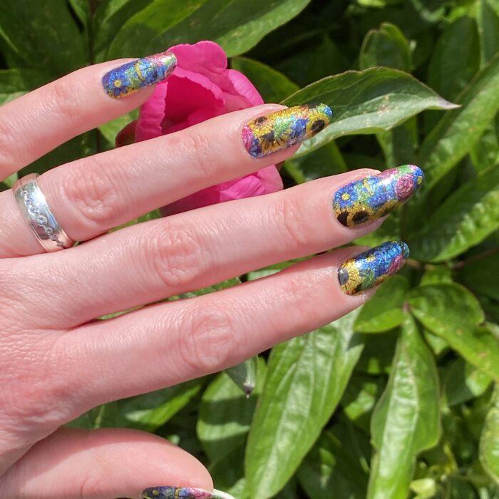 Bunte glitzernde Nagelfolie auf langen Nägeln vor einer pinken Blume im Sonnenlicht.